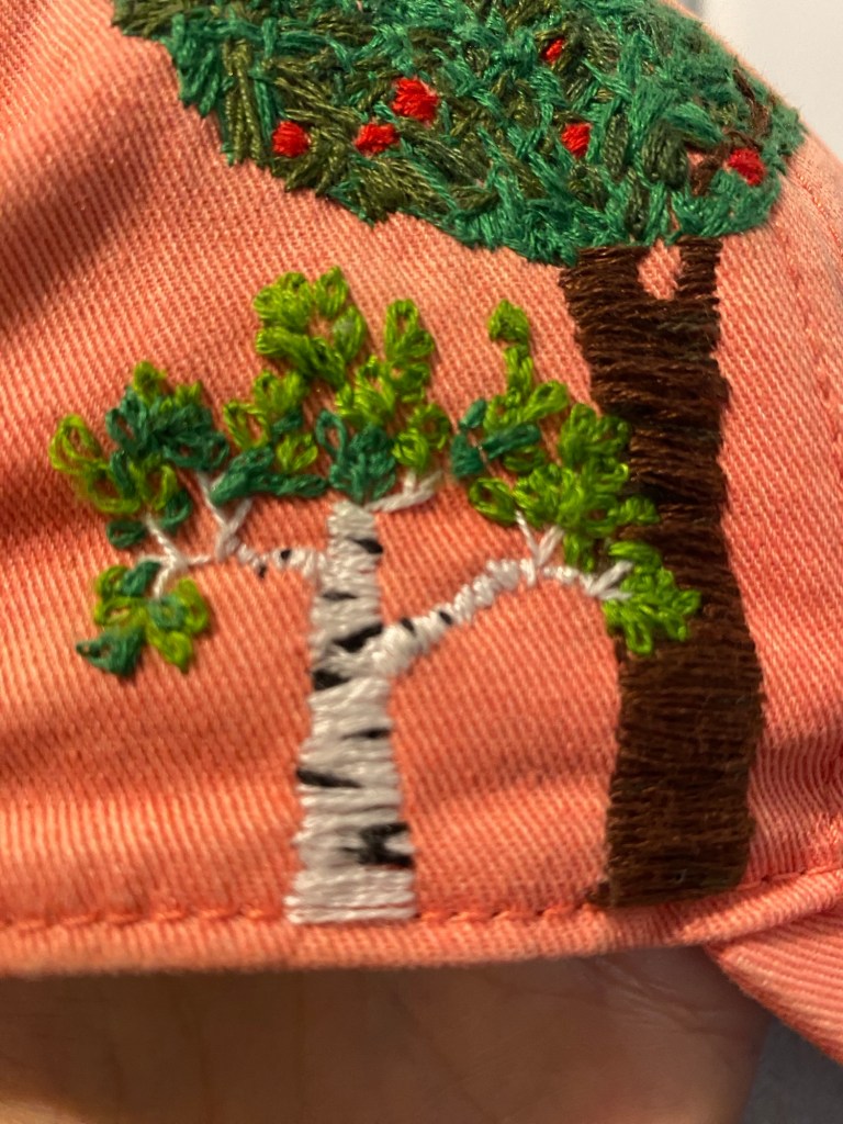 Kuvassa näkyy sivulta vaaleanpunainen lippis, johon on kirjottu valkorunkoinen koivu ja näkyy osa ruskearungoisesta vihreästä omenapuusta​ punaisilla omenoilla