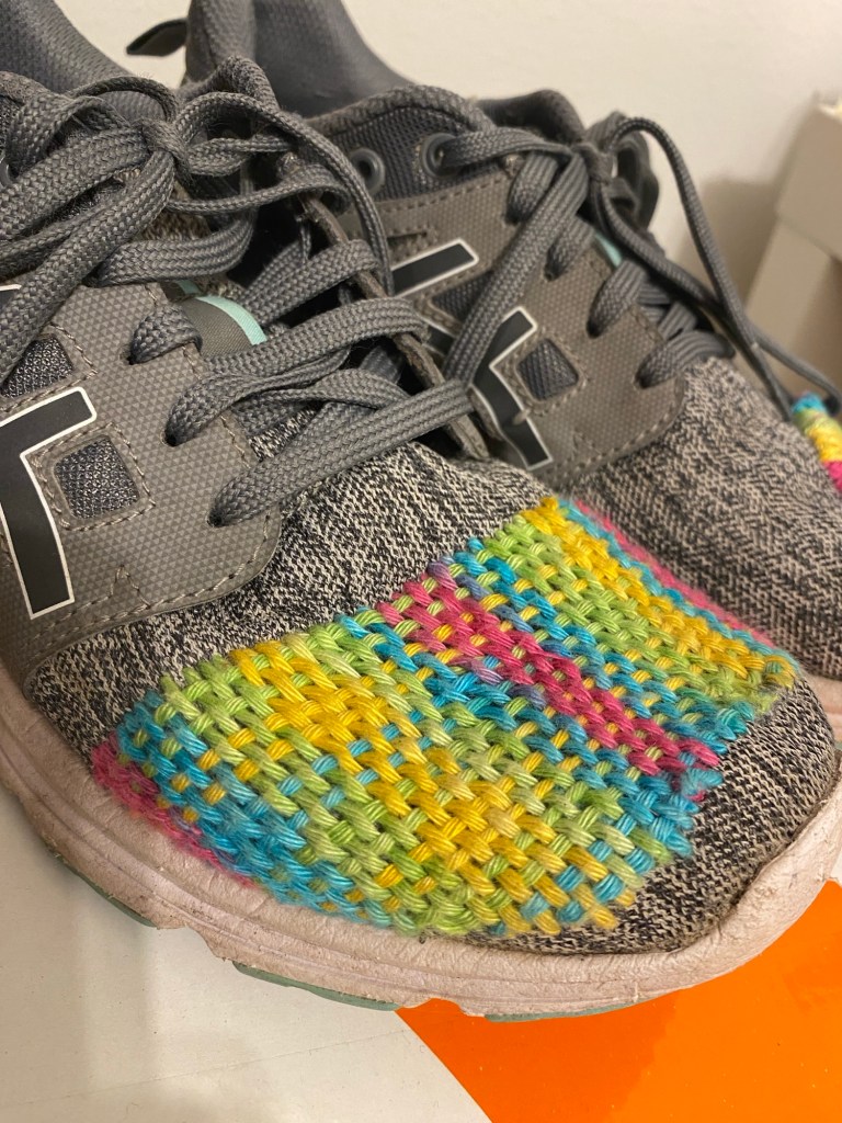 Kuvassa näkyy harmaat urheilukengät, jonka toisen etuosaan on ommeltu punomalla värikäs säännöllinen kuvio