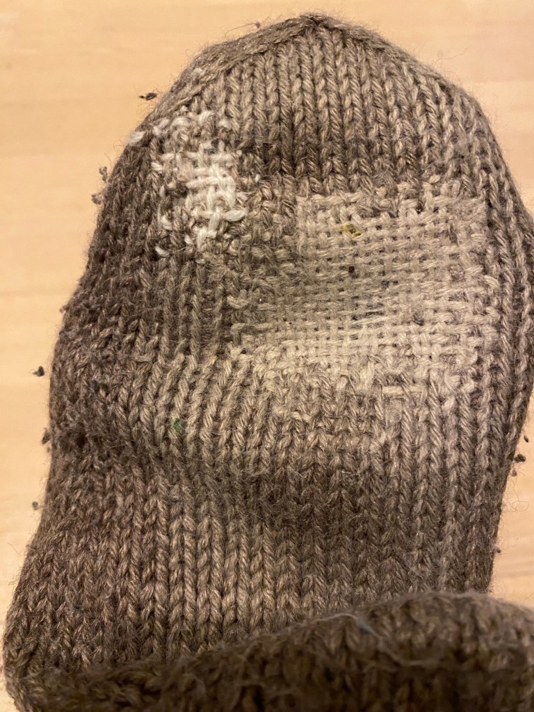 kuvassa harmaanruskea villa sukka, jota on paikattu valkoisella langalla kahdesta kohdasta parsimisen tavalla