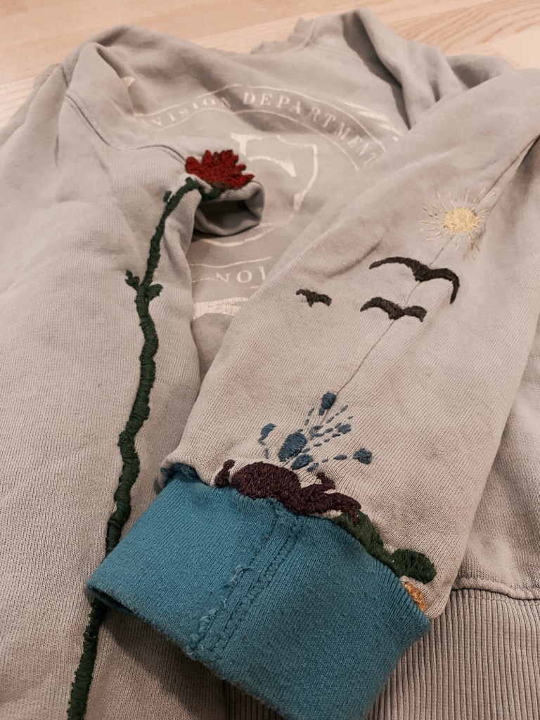 vaaleansinisen collegepaidan hihaa on korjattu kirjonnalla, jossa on meriläimiä (vettä roiskuttava mustekala, kilpikonna, kaloja) ja taivaalla lintujen siluetit