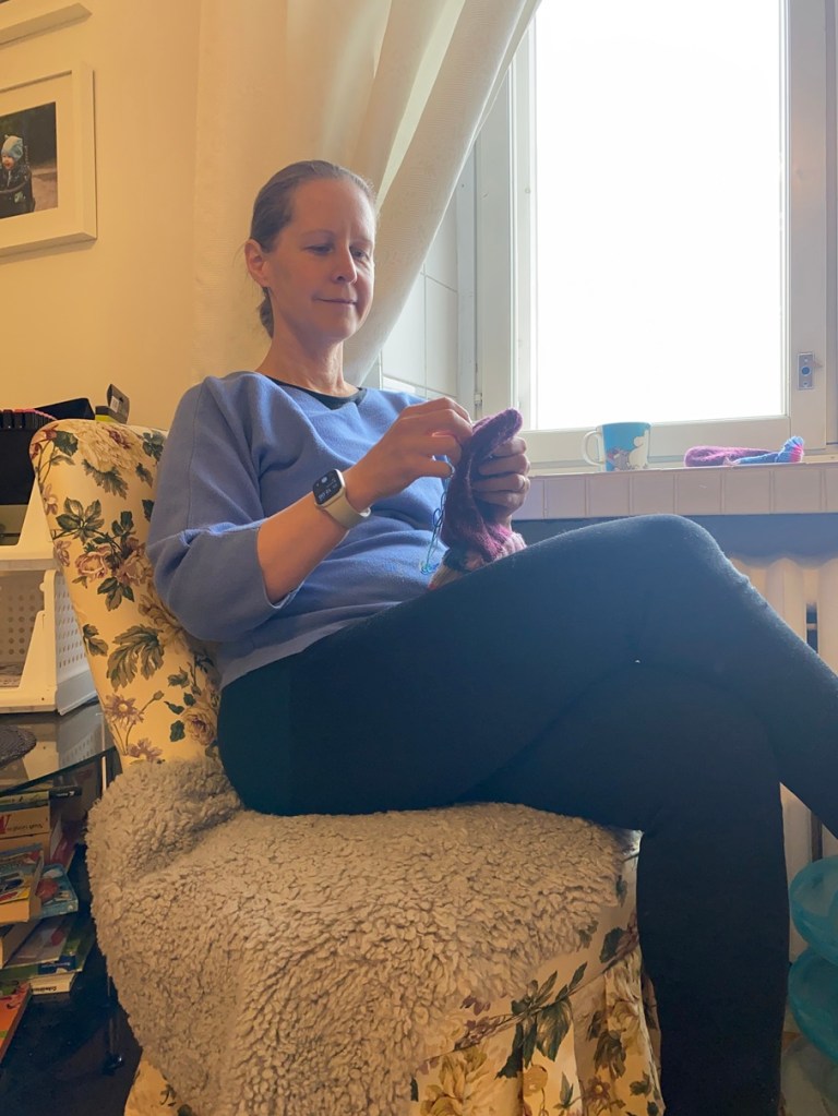 Nainen istuu ikkunan ääressä mukavassa kukkakuosisessa nojatuolissa ja paikkaa tyytyväisenä villasukan. Hänellä on sininen paita ja mustat housut. 