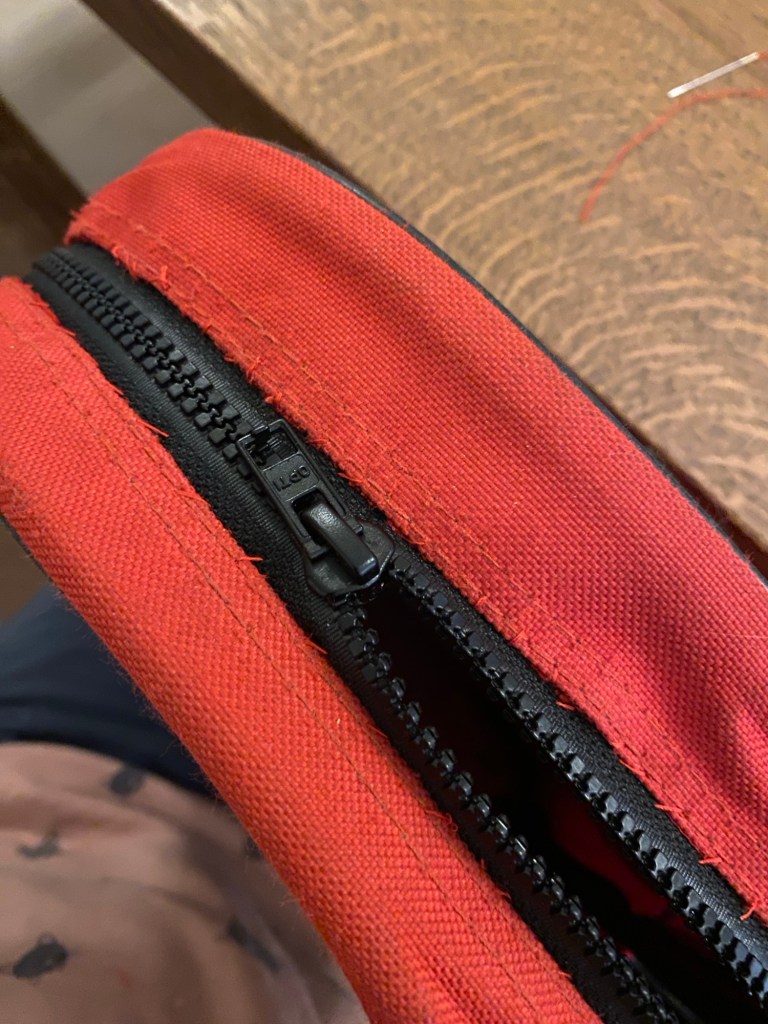 Näkyy punainen reppu päältäpäin, jossa on vahva musta vetoketju. Reunoista näkee, että vetoketju on ommeltu kiinni käsin punaisella langalla. 