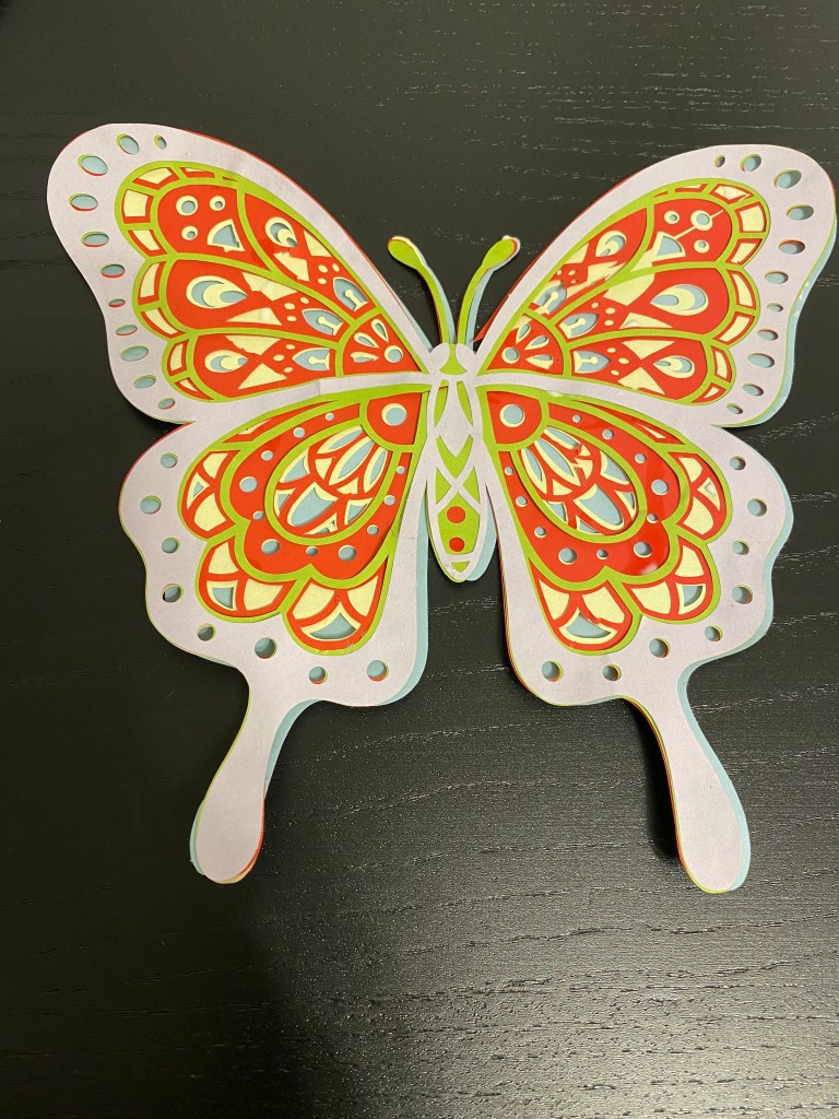 Värikäs ja koristeellinen paperinainen perhonen, jossa on punaisia, vihreitä ja keltaisia kuvioita, mustalla taustalla.