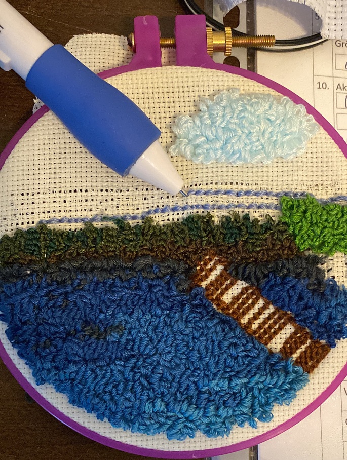 Työn alla ja keskeneräisenä tuftaustyö ympyrämuotoisessa violetissa raamissa. Tuftattuna on jo eri siniväreillä meri, jonka halki menee kirjottu ruskea laituri. Ylempänä ruskeaa ja vihreää kasvillisuutta. Tuftausneula törröttää taivaan kohdalla, joka on kesken. Taivaalla pehmeän pörröinen pilvi. 